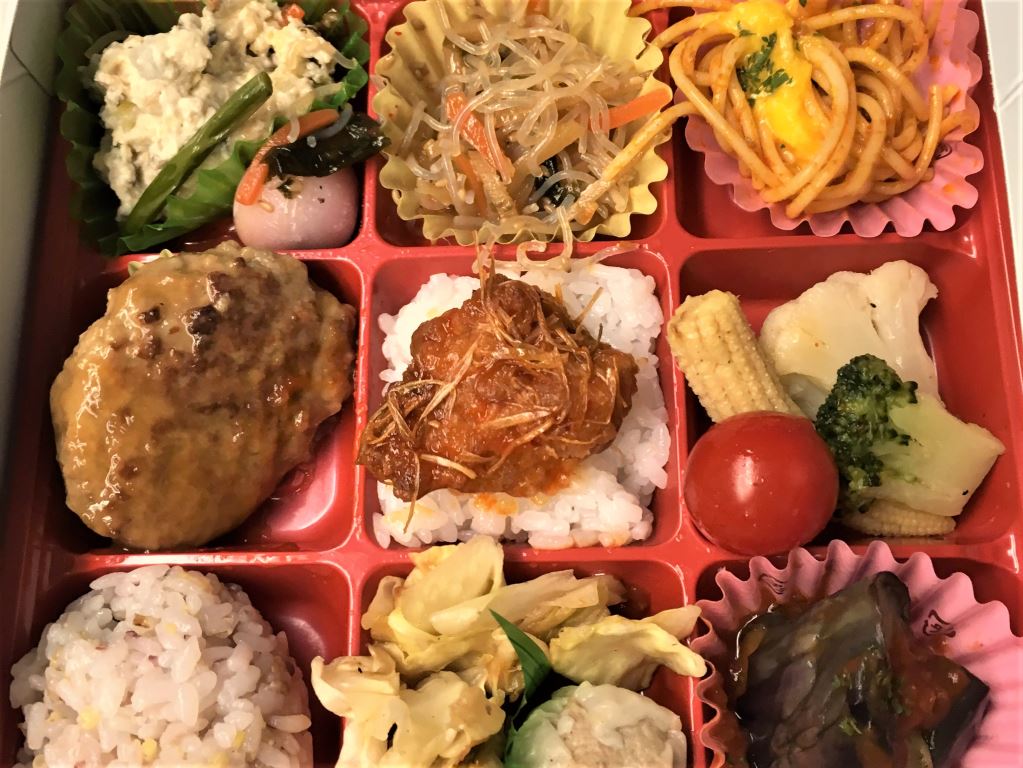 Ekiben - or train meal - in Japan. Nine small square compartments contain tiny portions of dishes including noodles, potato salad, vegetables and meat on rice.