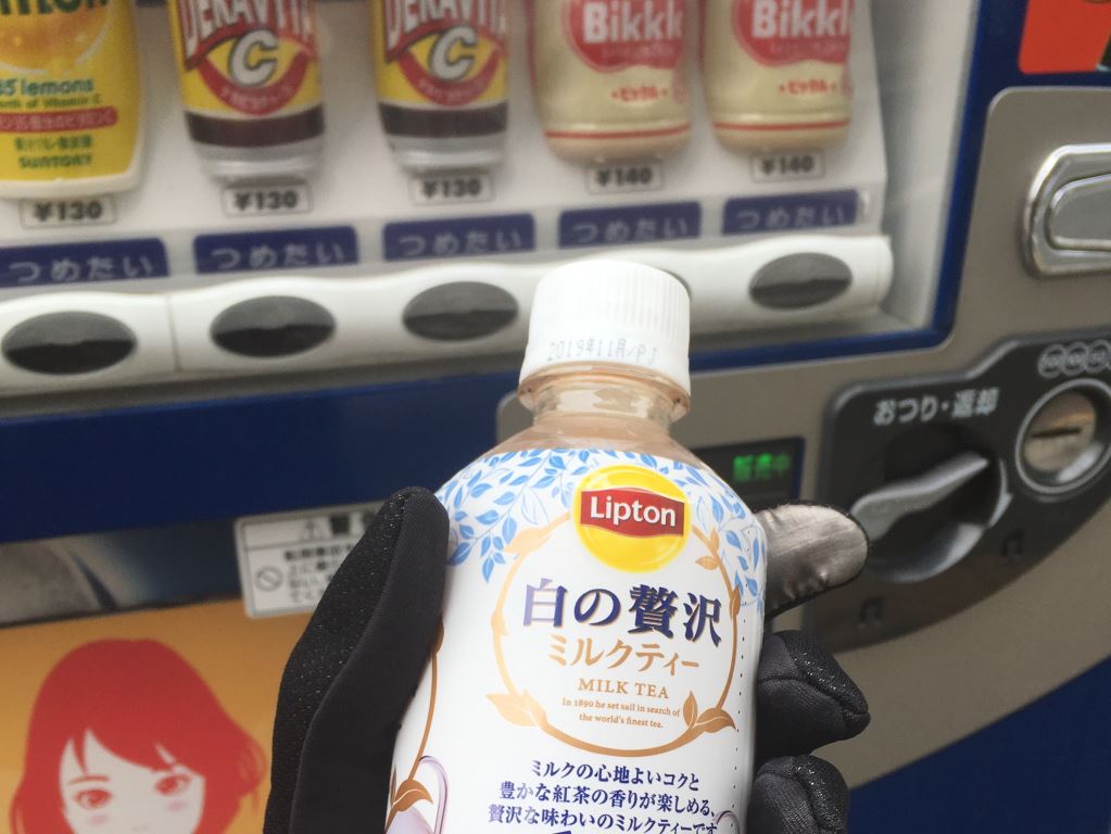 Hand wearing a black glove holds a bottle of hot Lipton Milk tea in Japan. Behind them is a vending machines selling different drinks. Hot vending machine drinks are one of the cool things to spot in Japan
