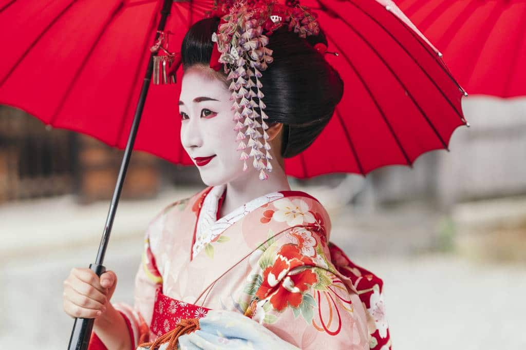 A geisha stands under a bright red umbrella. She has the traditional white face make up and red lipstick of a geisha - both lips are painted showing that she is a geisha not a maiko. Her kimono is salmon pink with red flowers