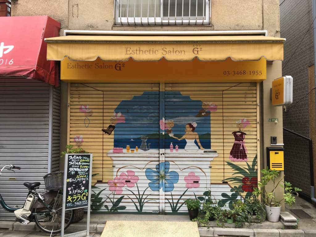 Shutter art in Shimokitazawa - seeing this is one thing to do in Tokyo in the morning. This art is on a beauty salon and it shows a woman sitting on a windowsill looking out to see with a cat. A shelf full of beauty products is behind her