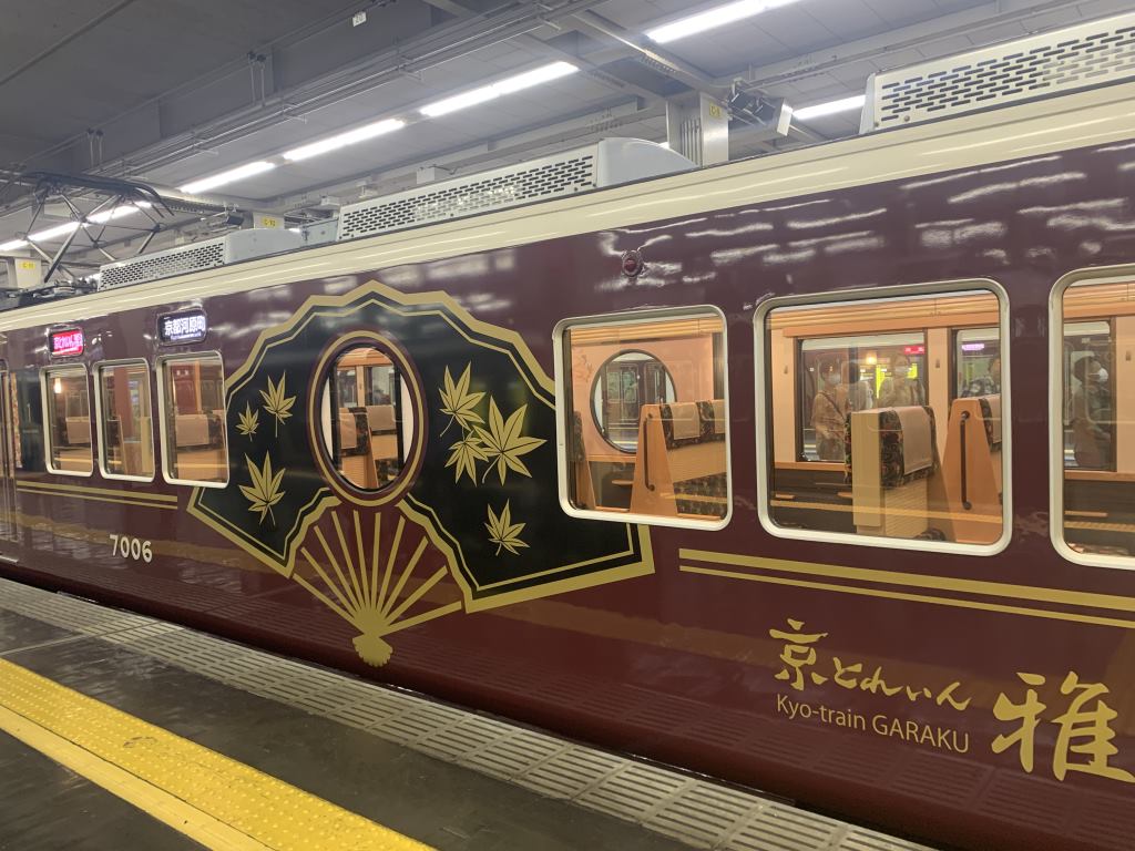 Exterior view of the Kyo Train Garaku between Osaka and Kyoto. It has an oxblood coloured carriage exterior with a black and gold fan drawing spanning the carriage.