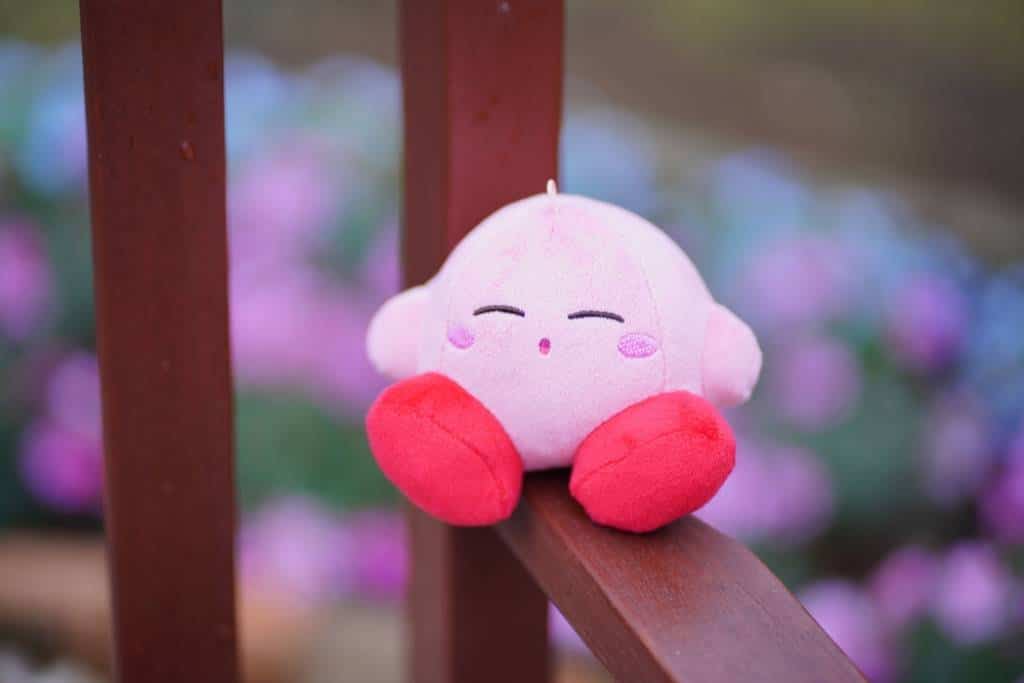 Stuffed Kirby toy sits on the side of a bench with flowers in the background