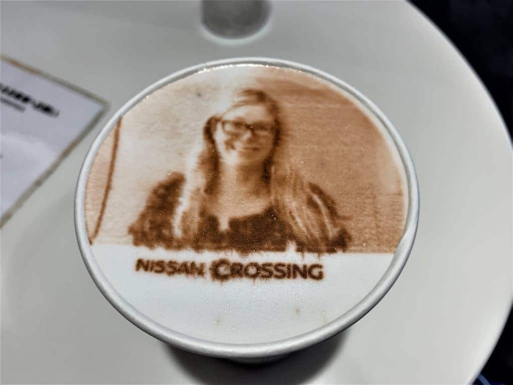 Cut of hot chocolate with a woman's face 'printed' on it in chocolate. The words Nissan Crossing are written on the top in powdered chocolate