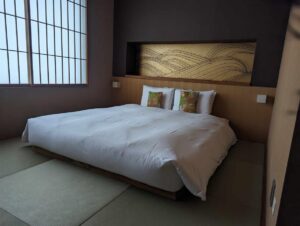 LArge double bed with a gold headboard in a room at the Misen hotel Kyoto. Paper screens over the windows give the room a traditional feel