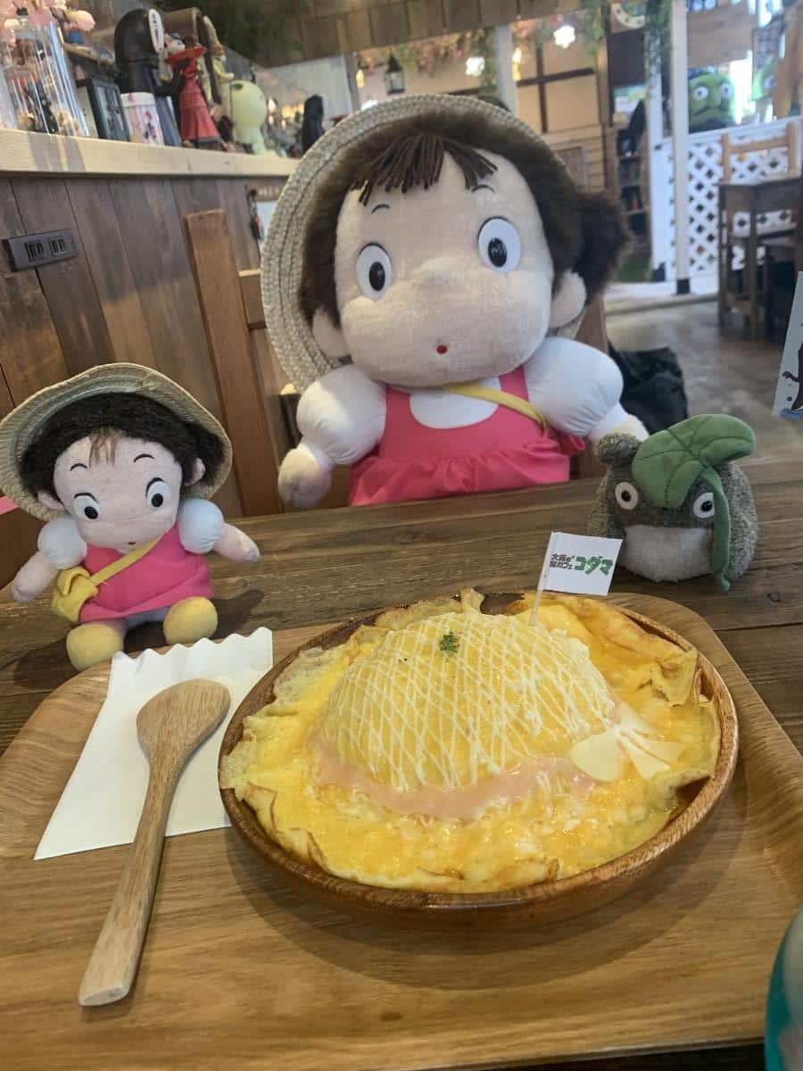 Omurice dish in Japan shaped like Mei's Hat from My Name is Totoro. Two dolls of Mei sits in the other chair on the seat and a small Totoro toy sits next to the omurice.
