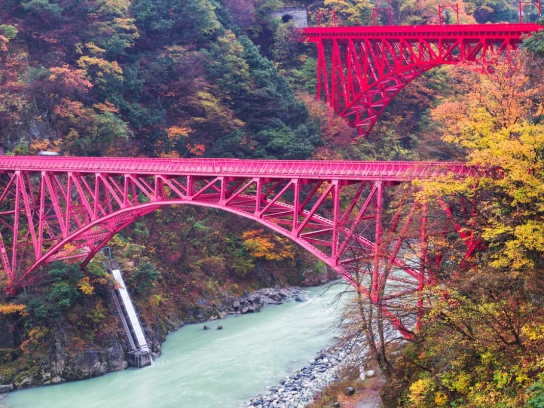 Two red bridges cross the blue waters of the Kurobe Gorge in Japan. There are pretty autumn leaves in the background.