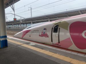 The pointy front of the Hello Kitty Shinkansen in the platform. The train is white with pink detailing. A picture of Hello Kitty in a train conductors outfit is on the side.