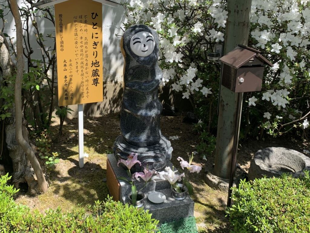 Black statue of a tall figure with a white smiling face. A sign behind it is in Japanese