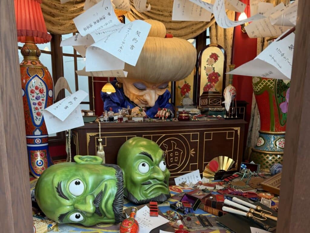 The witch from Spirited Away sits at a desk, the Kashira green heads are on the ground. 