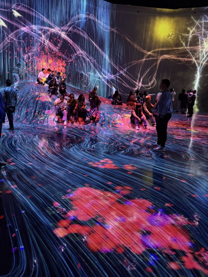 People sit on the raised 'rock' in the middle of teamLab Bordeless. Coloured lights and swirly lines are projected on the wall and floor around them. 