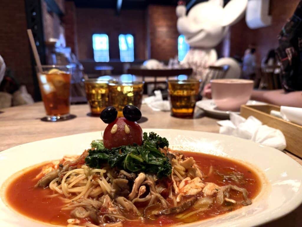 Pasta dish topped with tomato and mushroom sauce and kale. A tomato sits on top with two pine nuts and two olives to make it look like Mickey Mouse