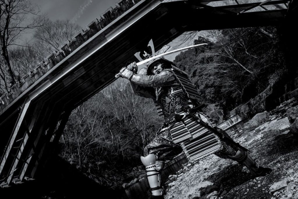 Men dressed in samurai armour wields a sword by a bridge in a black and white photo by Shogun Studio, Kyoto