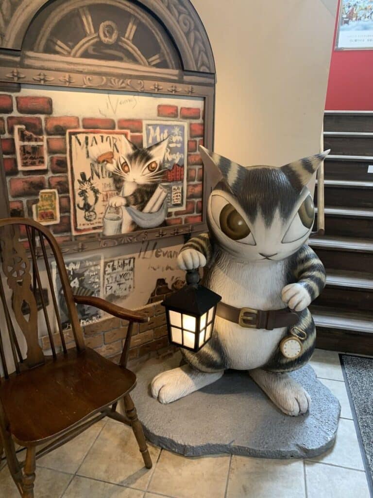 Model of Dayan the Cat in the museum. He is a grey and white stripy cat wearing a belt, a pocket watch (despite not having any pockets) and carrying a lantern.