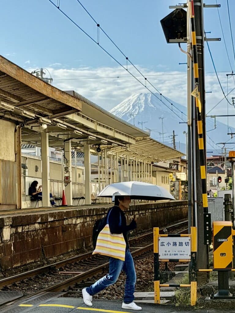 Man with white umbrella walks across a railway crossing by a station - Mount Fujhi is in the background. 