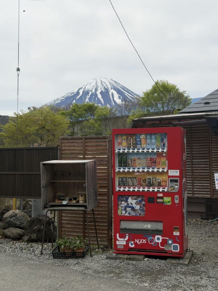A bright red vending machine sits in front of a wooden fence - Mount Fuji is behind it. 