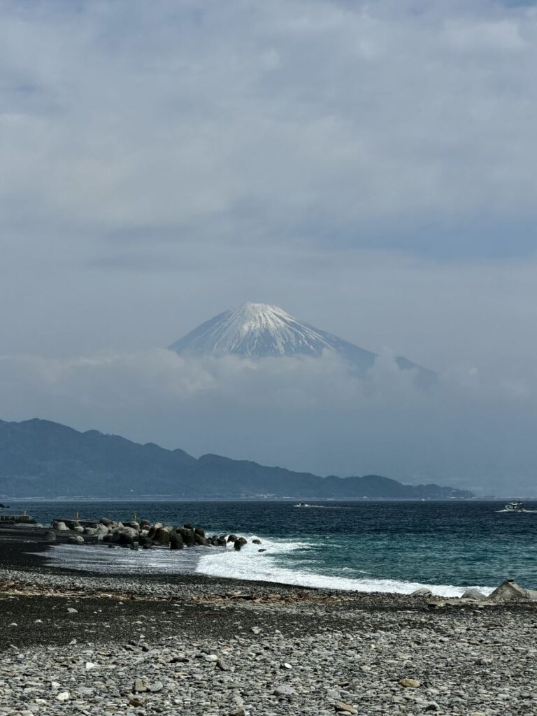 Mount Fuji's snow topped cone peeks out of the clouds - a pebbly black sand beach and sea are in the foreground of the picture